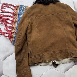 May include: A brown suede jacket with a black shearling collar. The jacket has a button closure and two front pockets. The interior is lined with a cream-colored, fuzzy material. The jacket is displayed on a patterned surface.