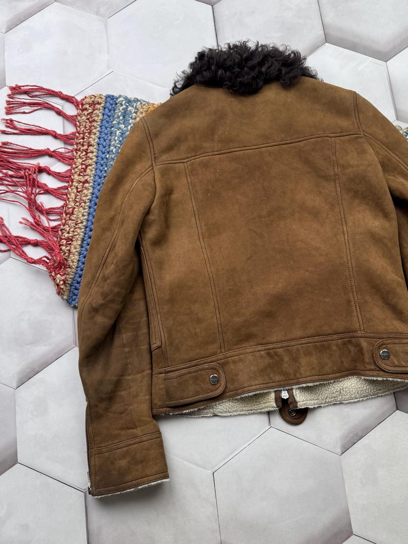 May include: A brown suede jacket with a black shearling collar. The jacket has a button closure and two front pockets. The interior is lined with a cream-colored, fuzzy material. The jacket is displayed on a patterned surface.