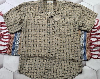 Könnte beinhalten: Kurzärmeliges Button-Down-Hemd mit einem sich wiederholenden Muster aus beigen und marineblauen Ovalen. Das Hemd hat einen Kragen und eine einzelne Brusttasche. Das Hemd liegt auf einem dekorativen Teppich mit rotem Fransen.