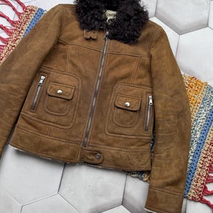 May include: A brown suede jacket with a black shearling collar. The jacket features a front zipper, two front pockets with button closures, and silver zippers on the pockets. The jacket has a classic design.