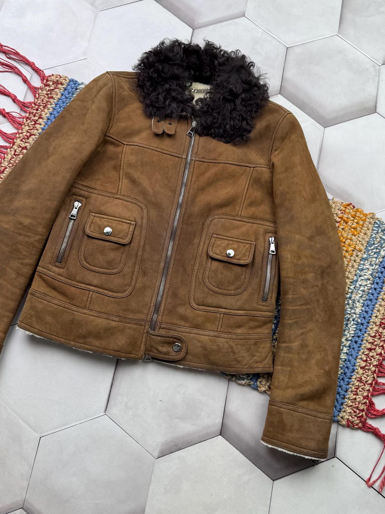 May include: A brown suede jacket with a black shearling collar. The jacket features a front zipper, two front pockets with button closures, and silver zippers on the pockets. The jacket has a classic design.