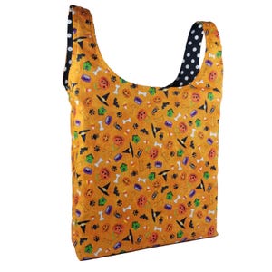 May include: Reversible Halloween-themed tote bag. One side features an orange background with Halloween icons like pumpkins, witch hats, and bones. The reverse side is black with white polka dots. The bag is designed for carrying items.