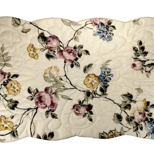 May include: A white quilted placemat with a floral pattern featuring pink, yellow, and blue flowers. The placemat has a scalloped edge.