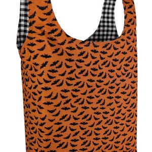 May include: Orange tote bag with a black bat pattern. The interior and handles are black and white gingham. The bag is made of fabric and is designed for carrying items.