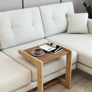 Pode incluir: Uma mesa lateral de madeira clara com uma bandeja embutida, com uma chávena e pires, uma revista e um livro. A mesa está posicionada ao lado de um sofá seccional cinza claro. A mesa tem um design simples e moderno.