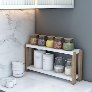 May include: A white two-tiered kitchen shelf with wooden supports, holding glass jars with various spices and white canisters labeled "FLOUR" and "SUGAR". Additional small white bowls and cups are on the countertop.