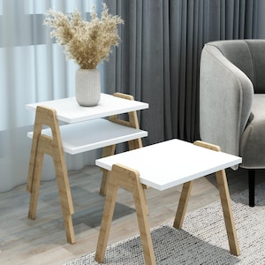 Può includere: Due tavolini bianchi con gambe in legno chiaro. Un tavolo ha due livelli, l'altro uno solo. Un vaso beige con fiori secchi è posto sul livello superiore. I tavoli sono su un pavimento in legno chiaro con un tappeto grigio e bianco.