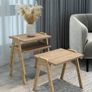 May include: Two light-colored wooden side tables with a natural finish. One table has two tiers, and the other is a single-tier table. A vase with dried flowers sits on the top tier of the two-tiered table. The tables are on a patterned rug.