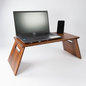 Può includere: Un supporto per laptop in legno con finitura marrone scuro. Il supporto ha due gambe pieghevoli e un supporto per telefono integrato. Un laptop e uno smartphone sono posizionati sul supporto.