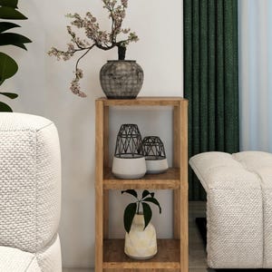 May include: A wooden shelving unit with decorative items. A bonsai tree in a textured grey pot sits on top. Below are two black wire lanterns over white bases, and a vase with a plant. The shelf is in a room with a green curtain.