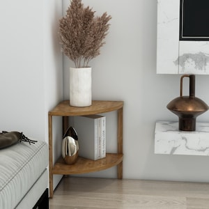May include: A wooden corner shelf unit with two levels. A white vase holds dried brown plants. A silver decorative object and a book are on the lower shelf. The room has a modern aesthetic with a white wall and a marble-like accent.