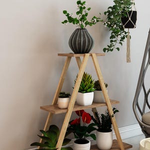 Może przedstawiać: Drewniany stojak na rośliny z trzema półkami, na których znajdują się różne rośliny doniczkowe i dekoracyjny wazon. Widoczny jest również wiszący kwietnik z makramy i szary doniczek. Stojak ma trójkątny kształt.