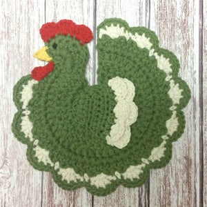 May include: Green crocheted chicken kitchen dishcloth with a white scalloped edge and a red comb.