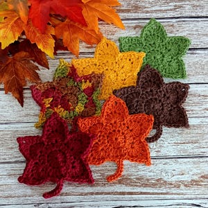 May include: Five crocheted leaf-shaped coasters in various fall colors, including red, orange, yellow, brown, and green. The coasters are arranged on a white wooden surface.