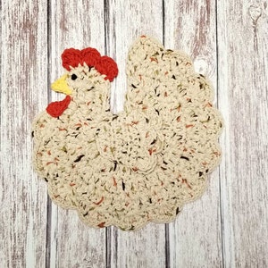 May include: A crocheted kitchen dishcloth shaped like a chicken. The chicken is beige with red and yellow accents. The dishcloth is on a white wooden background.