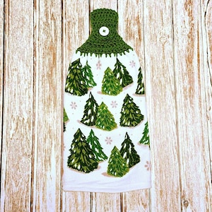 May include: A white kitchen towel with a green crocheted top and button closure. The towel features a pattern of green Christmas trees and pink snowflakes. The towel is designed to hang from a hook.