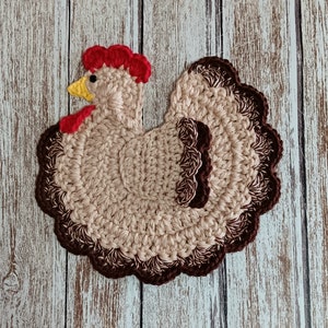 May include: A crocheted brown and beige chicken with a red comb and a yellow beak. The chicken is on a white wooden background.