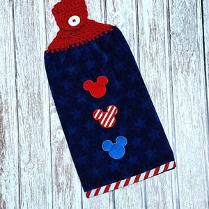 Puede incluir: Una toalla de cocina colgante azul marino con una parte superior de ganchillo roja y un bot&oacute;n blanco. La toalla tiene un patr&oacute;n de estrellas sutil y tres apliques con forma de rat&oacute;n en rojo, rayas rojas y blancas, y azul. Borde a rayas.