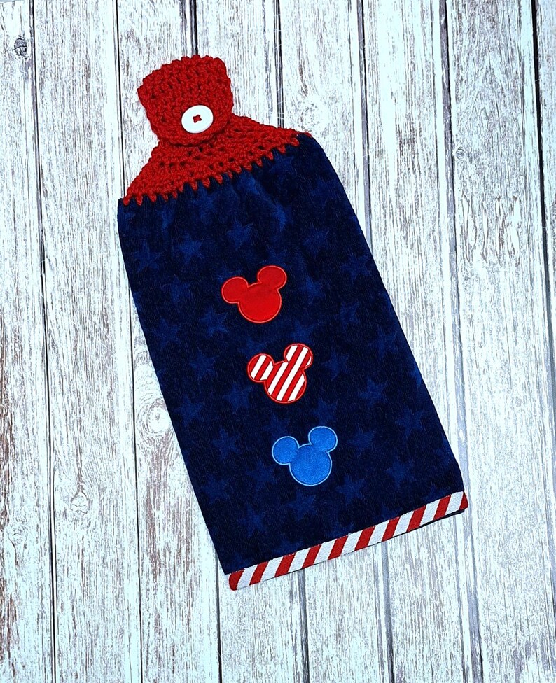 Puede incluir: Una toalla de cocina colgante azul marino con una parte superior de ganchillo roja y un bot&oacute;n blanco. La toalla tiene un patr&oacute;n de estrellas sutil y tres apliques con forma de rat&oacute;n en rojo, rayas rojas y blancas, y azul. Borde a rayas.