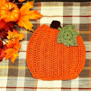 May include: A crocheted orange pumpkin with a green crocheted leaf on a plaid fabric background.