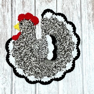 May include: A crocheted black and white chicken with a red comb and yellow beak. The chicken is sitting on a white wooden background.