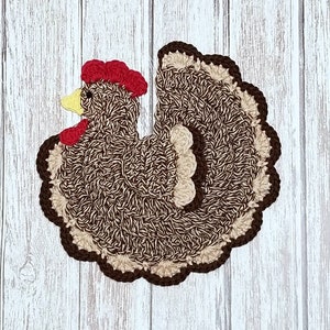 May include: A handmade crochet chicken-shaped trivet. The trivet features a brown and beige body with a red comb and wattle, and a yellow beak. The edges are trimmed in dark brown. The trivet is placed on a white wooden surface.