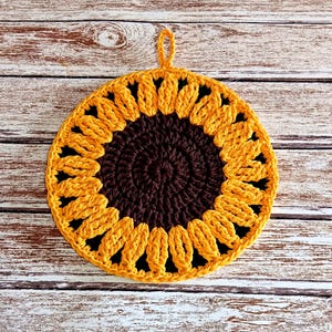 May include: A handmade, round crochet potholder in the shape of a sunflower. The center is dark brown, surrounded by yellow petals. A small loop is attached for hanging. The potholder is placed on a light-colored wooden surface.
