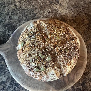 May include: A round loaf of sourdough bread with a variety of seeds on top, including pumpkin seeds, sesame seeds, and flax seeds. The bread is sitting on a wooden cutting board.