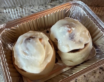 May include: Two frosted cinnamon rolls in a foil container. The rolls are light brown with a swirl of cinnamon and a thick layer of white frosting. Other containers of cinnamon rolls are visible in the background.
