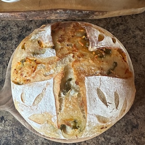 May include: A golden brown sourdough bread loaf with a crusty exterior and a soft, airy interior. The bread is topped with green peppers and cheese, and has a decorative leaf pattern on the top.