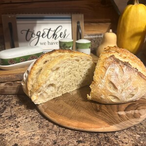 K&ouml;nnte beinhalten: Eine Nahaufnahme eines frisch gebackenen Brotlaibs, der in zwei H&auml;lften geschnitten ist, auf einem Holzbrett. Das Brot hat eine hellbraune Kruste und ein weiches, wei&szlig;es Inneres. Das Brot liegt auf einem Holzbrett mit rustikaler brauner Oberfl&auml;che.