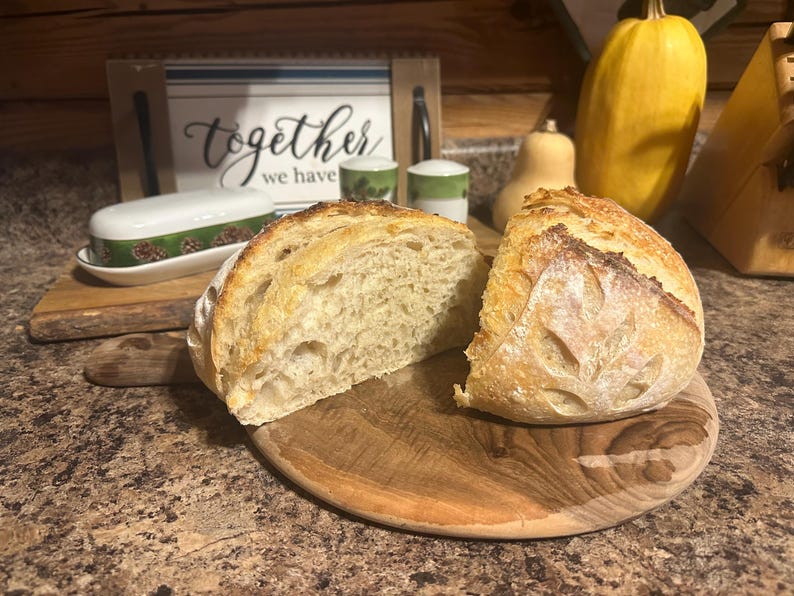 K&ouml;nnte beinhalten: Eine Nahaufnahme eines frisch gebackenen Brotlaibs, der in zwei H&auml;lften geschnitten ist, auf einem Holzbrett. Das Brot hat eine hellbraune Kruste und ein weiches, wei&szlig;es Inneres. Das Brot liegt auf einem Holzbrett mit rustikaler brauner Oberfl&auml;che.