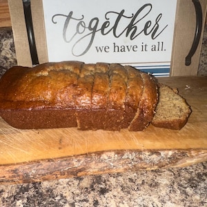 May include: A sliced loaf of banana bread on a wooden cutting board. The bread is golden brown with a slightly textured crust. A slice is cut off and placed next to the loaf. A sign in the background reads "Together we have it all."