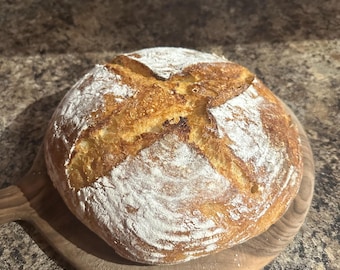 May include: A freshly baked loaf of sourdough bread with a crispy crust and a soft, airy interior. The bread is scored with a cross on top and dusted with flour.