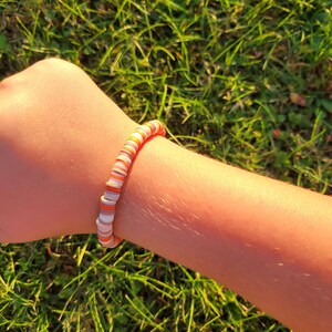 May include: A bracelet made of small, round, colourful beads. The beads are orange, white and brown, and they are arranged in a striped pattern. The bracelet is worn on a person's wrist.