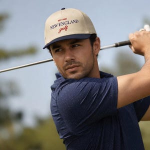 May include: A beige and navy blue baseball cap with the text "NEW ENGLAND golf" in red script. The cap is worn by a person holding a golf club, with a blue shirt.