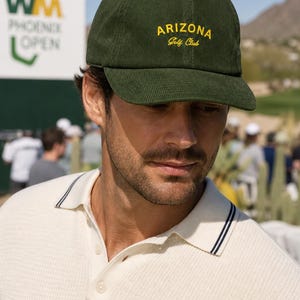 May include: A green corduroy baseball cap with "ARIZONA Golf Club" embroidered in yellow. The cap is worn by a person wearing a white polo shirt with navy blue trim. The background shows a golf course and a sign for the WM Phoenix Open.