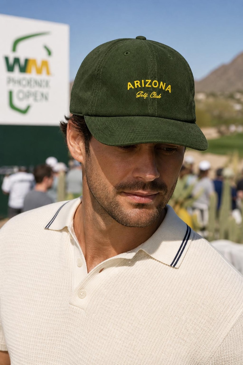 May include: A green corduroy baseball cap with "ARIZONA Golf Club" embroidered in yellow. The cap is worn by a person wearing a white polo shirt with navy blue trim. The background shows a golf course and a sign for the WM Phoenix Open.