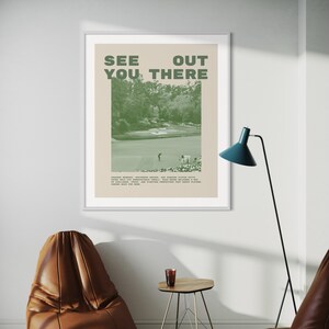 a framed poster with the text "SEE YOU THERE" displayed on a wall, along with a golf course image. There is a brown leather chair and a small table in the foreground, and a blue floor lamp is visible in the background.