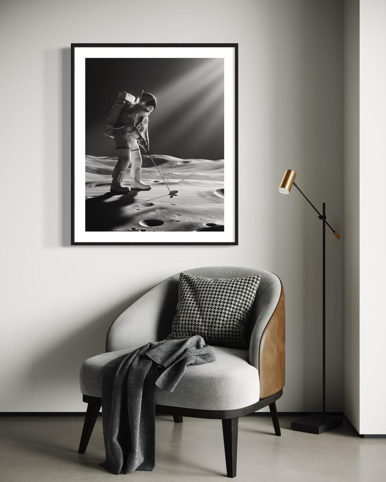 May include: Black and white photograph of an astronaut playing golf on the moon. The astronaut is wearing a spacesuit and carrying a golf club. Sunlight beams down on the astronaut. The photo is displayed in a black frame on a wall above a gray armchair with a brown leather side. A gold floor lamp is in the corner of the room.