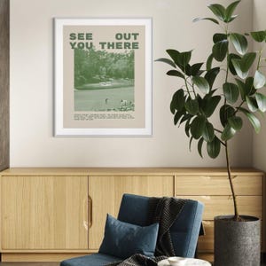 a framed poster with the text "See You There" hanging on a wall above a wooden cabinet and a blue armchair in a living room setting.