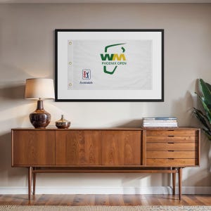 May include: A framed white flag with the text "WM Phoenix Open" in green and yellow, with the city "Scottsdale" below. The flag is mounted on a wall above a mid-century modern wooden cabinet with a lamp and decorative items.
