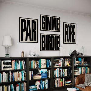 May include: Four framed wall art pieces with black text on a cream background. The words "PAR", "GIMME", "BIRDIE", and "FORE" are displayed. The art hangs above a dark bookshelf filled with books, a lamp, and decorative items.