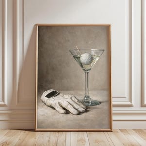 May include: A framed print featuring a golf ball in a martini glass with a toothpick and a white golf glove. The artwork has a neutral color palette with a vintage aesthetic.
