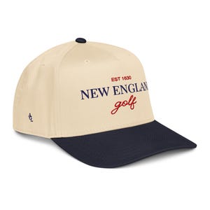 May include: A cream-colored baseball cap with a navy blue brim. The cap features the text "NEW ENGLAND golf" in navy blue and red, with "EST 1630" above. A small logo is on the side.