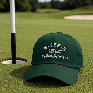 Pode incluir: Boné de beisebol verde escuro com texto branco "6-TEE-5 BIRTHDAY CLASSIC Still On Par". O boné está em um campo de golfe verde, perto de um buraco e uma bandeira.