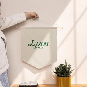 May include: A cream-colored banner with the name "Liam James" in green script. The banner is suspended from a wooden rod and string. A potted plant in a yellow container sits below the banner.