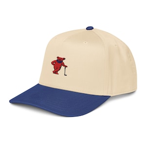 Könnte beinhalten: Baseballcap mit cremefarbener Krone und marineblauem Schirm. Die Kappe hat einen gestickten roten Bären, der einen Hockeyschläger hält. Die Kappe hat einen gebogenen Schirm und einen Knopf oben.
