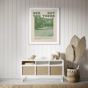 a framed poster with the text "See You There" hanging on a wall above a white cabinet with three shelves. The cabinet has a woven design and is positioned next to a potted plant with large leaves.