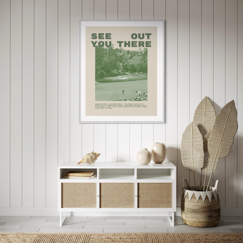 a framed poster with the text "See You There" hanging on a wall above a white cabinet with three shelves. The cabinet has a woven design and is positioned next to a potted plant with large leaves.